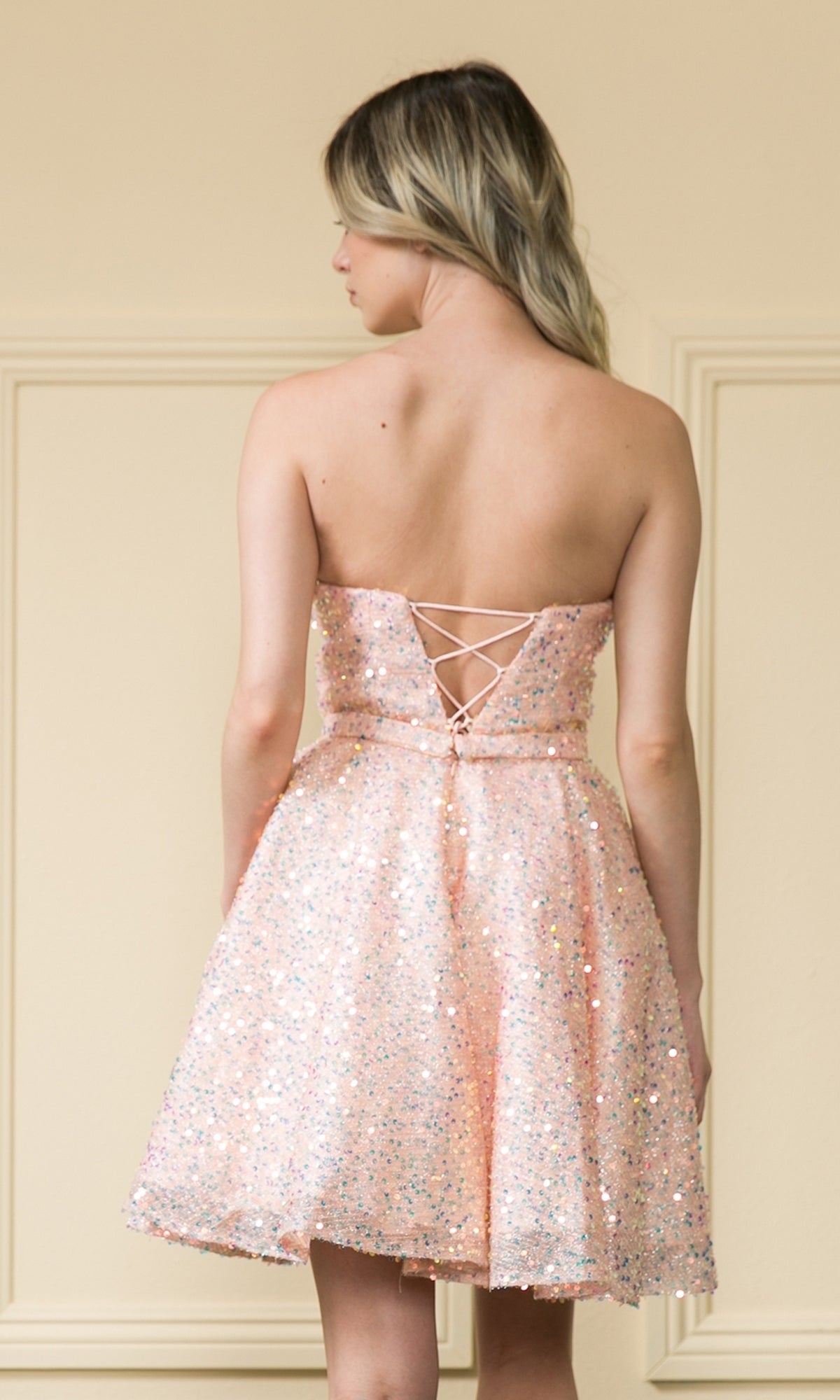 A woman in the Short A-Line Sequin Homecoming Dress 8930, a sparkly light pink strapless style with a lace-up back, stands facing away from the camera against a beige wall.